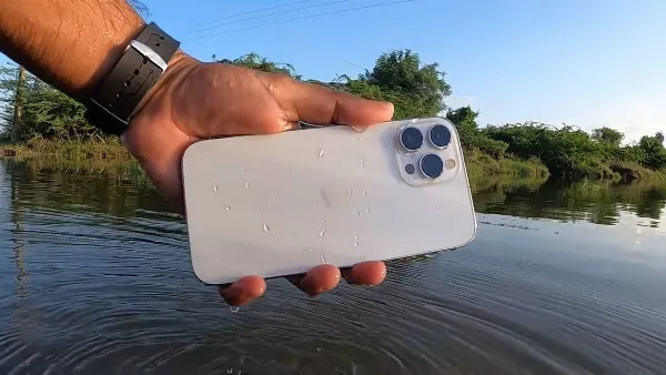 Một chiếc iPhone 13 màu xanh dương đang được cầm trên tay, nền phía sau là một bể cá với nhiều loại cá cảnh đầy màu sắc, minh họa cho khả năng chống nước của iPhone 13